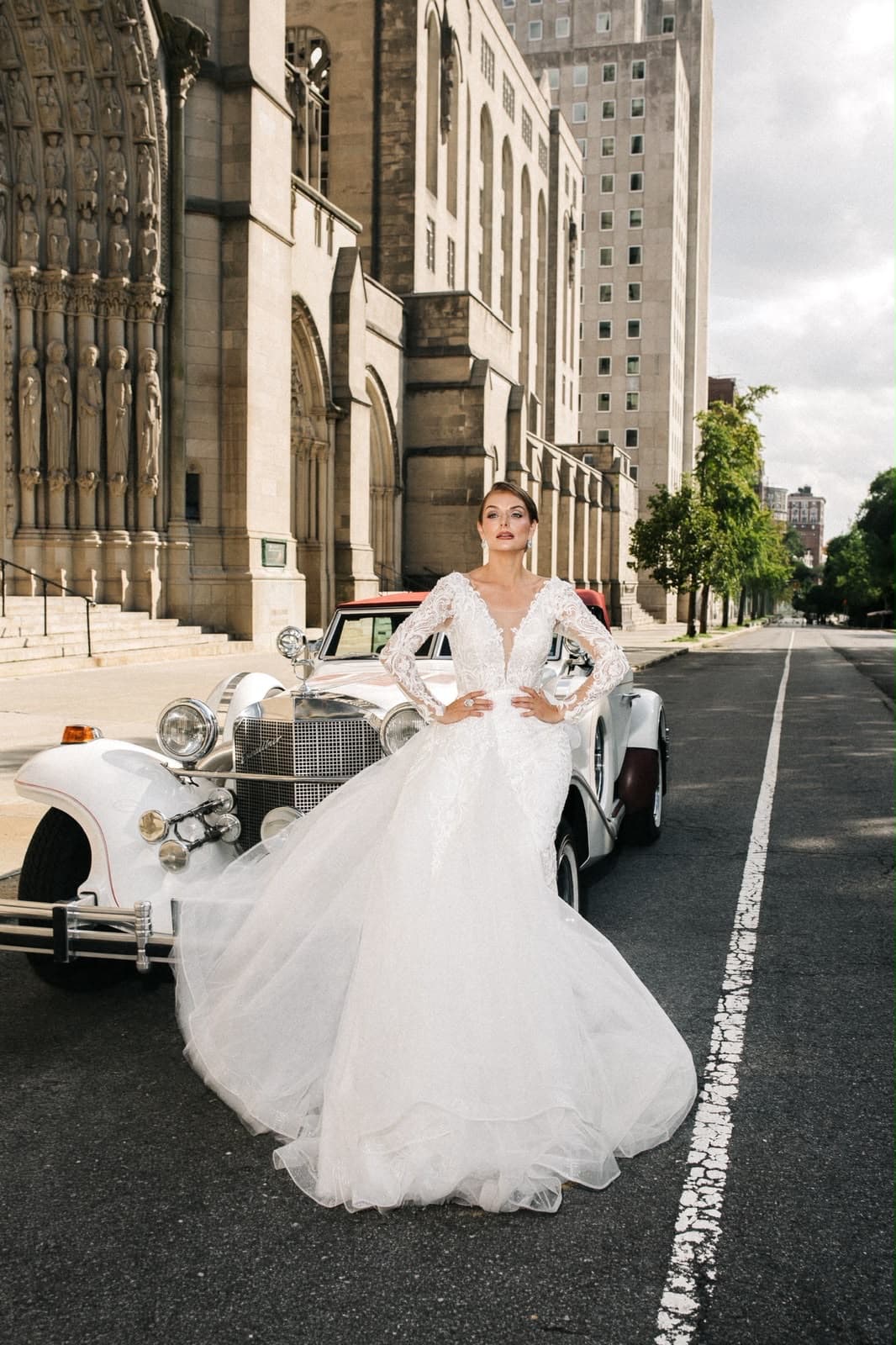 Bride in a lace gown beside a vintage car at a New York cathedral, bridal makeup by Malaz
